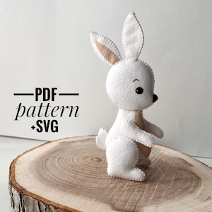 May include: A white felt bunny with brown felt ears and black eyes. The bunny is standing on a wooden surface. The text "PDF pattern +SVG" is written in black on a white background.