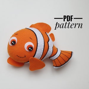 Pode incluir: Um peixe-palhaço de feltro laranja e branco com olhos pretos e contorno preto. O peixe está deitado sobre uma superfície branca. O texto "PDF pattern" está escrito em preto acima do peixe.