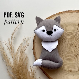 Può includere: Un lupo di feltro grigio con dettagli in feltro bianco, seduto su una fetta di legno. Il lupo ha il naso nero e gli occhi neri. Il testo "PDF, SVG pattern" è scritto in nero su sfondo bianco.