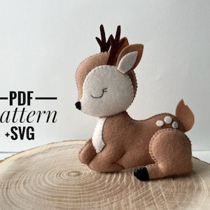 Puede incluir: Un ciervo de fieltro marrón con manchas blancas y los ojos cerrados. El ciervo tiene cuernos y está acostado sobre una superficie de madera. El texto "PDF pattern +SVG" está en negro sobre fondo blanco.
