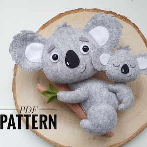 May include: A gray felt koala holding a smaller gray felt koala. Both koalas have white felt ears and black felt noses. The larger koala has black eyes and a smile. The smaller koala has closed eyes. The koalas are sitting on a wooden surface. The text "PDF PATTERN" is written below the koalas.