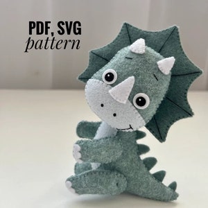 Può includere: Un giocattolo di peluche di triceratopo in feltro verde con dettagli bianchi. Il giocattolo ha occhi neri e un sorriso cucito. Il giocattolo è seduto su una superficie bianca. Modello PDF, SVG.