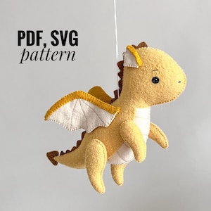 May include: A felt dragon toy with yellow wings and a brown head. The dragon is hanging from a white string. The text "PDF, SVG pattern" is written in black on a white background.