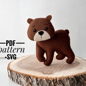 Puede incluir: Un oso de fieltro marrón con ojos negros y nariz blanca. El oso está de pie sobre una rodaja de madera. El texto "PDF pattern + SVG" está escrito en negro sobre fondo blanco.