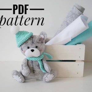 May include: A gray felt teddy bear wearing a light green crocheted hat and scarf. The bear is sitting in a white wooden crate with felt fabric in the background. The text "PDF pattern" is written above the bear.