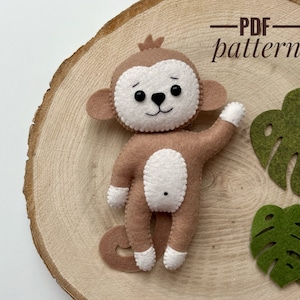May include: A brown felt monkey toy with white felt accents and black eyes. The monkey is holding its arm up in the air. The toy is sitting on a wooden surface with a green felt leaf behind it. The text "PDF pattern" is written in black on a white background above the monkey.