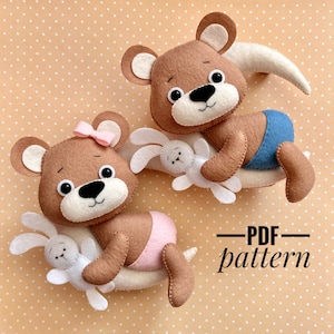 Könnte beinhalten: Zwei Teddybären aus Filz, die weiße Filzhasen halten. Die Bären sitzen auf einer Halbmondform. Der Bär links trägt eine rosa Windel und eine rosa Schleife. Der Bär rechts trägt eine blaue Windel. Der Text "PDF pattern" steht unter den Bären.