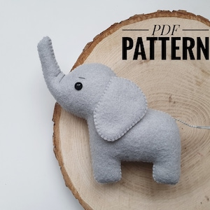 May include: A gray felt elephant plush toy with black eyes. The toy is sitting on a wooden slice with the text "PDF PATTERN" written on it.