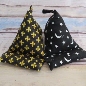 May include: Two triangular fabric phone stands. One is black with gold fleur-de-lis patterns. The other is black with white crescent moons and stars. Both have a slot to hold a phone.