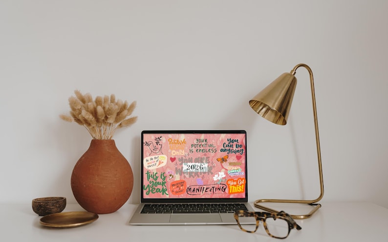 K&ouml;nnte beinhalten: Ein Laptop zeigt einen rosa Hintergrund mit inspirierendem Text und Grafiken. Eine goldene Schreibtischlampe, eine Terrakotta-Vase mit getrockneten Blumen und eine Brille stehen auf dem wei&szlig;en Schreibtisch. Eine kleine Holzschale und ein goldfarbener Teller sind ebenfalls vorhanden.