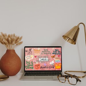 K&ouml;nnte beinhalten: Ein Laptop zeigt einen rosa Hintergrund mit inspirierendem Text und Grafiken. Eine goldene Schreibtischlampe, eine Terrakotta-Vase mit getrockneten Blumen und eine Brille stehen auf dem wei&szlig;en Schreibtisch. Eine kleine Holzschale und ein goldfarbener Teller sind ebenfalls vorhanden.