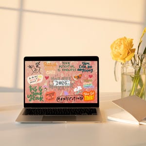 K&ouml;nnte beinhalten: Ein Laptop zeigt eine motivierende Grafik mit Text wie "This is your year" und "Manifesting This!". Ein Glas, eine Vase mit gelben Blumen und ein Notizbuch liegen auf einem wei&szlig;en Schreibtisch.