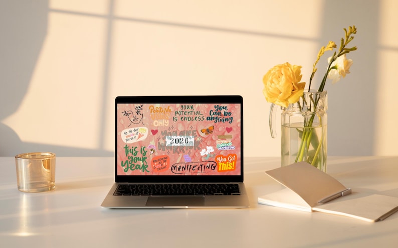 K&ouml;nnte beinhalten: Ein Laptop zeigt eine motivierende Grafik mit Text wie "This is your year" und "Manifesting This!". Ein Glas, eine Vase mit gelben Blumen und ein Notizbuch liegen auf einem wei&szlig;en Schreibtisch.