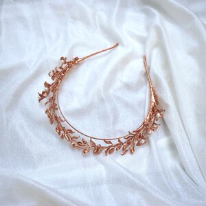 May include: A copper-coloured bridal headband with a delicate leaf and pearl design. The metal headband features intricate detailing of leaves and small, round pearls. The headpiece is set against a white, textured background.