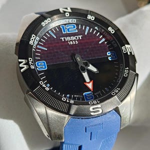 May include: A Tissot watch with a black face, blue numerals, and a blue rubber strap. The watch has a stainless steel case and features the text "TISSOT 1853". The watch face displays various functions and measurements. The strap is a vibrant blue colour.