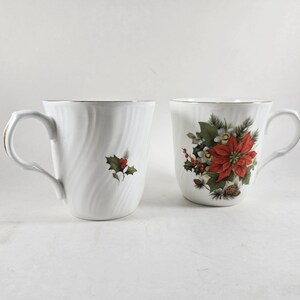 May include: Two white ceramic mugs with gold trim. One mug features a holly design, the other a poinsettia and pinecone arrangement. Both mugs have a curved handle.
