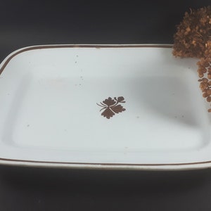 May include: Rectangular white ceramic serving platter with a brown border and a brown floral design in the center. The platter has rounded corners and is set against a dark background.