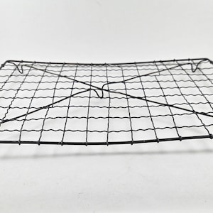 May include: A rectangular black wire cooling rack featuring a grid pattern constructed from thin metal wires. The design includes two diagonal support wires that cross in the centre, providing structural stability to the flat, open-weave surface.