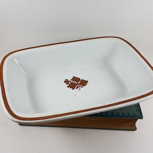 May include: A rectangular white enamel dish with a brown border and a brown leaf motif in the centre. The dish is placed on a vintage book with a green cover.