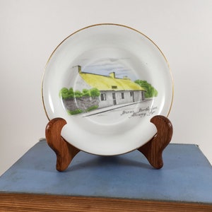 May include: A white decorative plate with a gold rim, featuring a painted scene of a cottage with a yellow thatched roof and the text "Burns Birthplace Alloway Ayr". The plate is displayed on a wooden stand.
