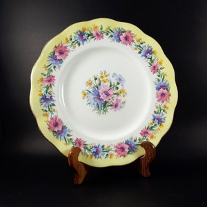 May include: A round ceramic plate with a scalloped edge and a pale yellow border. The plate features a central bouquet and a circular wreath pattern of pink, blue, and yellow flowers on a white background, finished with a gold-toned rim.