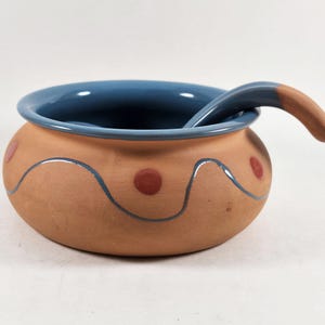 May include: A round terracotta bowl with a smooth matte finish, featuring a blue glazed interior and rim. The exterior is decorated with a hand-painted blue wavy line and small red dots. A matching terracotta ladle with a blue glazed handle rests inside.