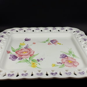Peut inclure: Un plat de service carré blanc avec un motif floral. Le plat présente un bord festonné avec des détails découpés et est décoré de fleurs roses, violettes et jaunes. Les fleurs sont peintes avec des feuilles vertes.