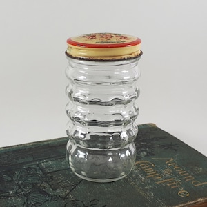 May include: A clear glass spice jar with a ribbed body and a metal lid. The lid is red and yellow with the word "Pepper" printed on it. The jar is on a vintage book.