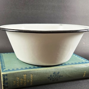 May include: A white enamel bowl with a black rim, placed on a vintage book with a teal cover. The bowl has a classic, flared shape. The book's cover features gold lettering and decorative patterns, enhancing the vintage aesthetic.