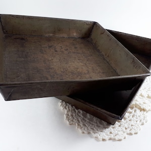 May include: Two vintage square baking trays with a well-worn, aged appearance. The trays are made of metal and have a dark, brownish-grey colour. They are stacked slightly, revealing their depth and shape. The trays sit on a white surface with a lace doily.