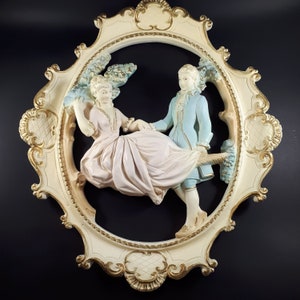 May include: Cream-colored wall decor featuring a romantic scene of a couple in period clothing. The oval frame has gold accents. The man wears a light blue coat, and the woman wears a pink dress. The background is black.