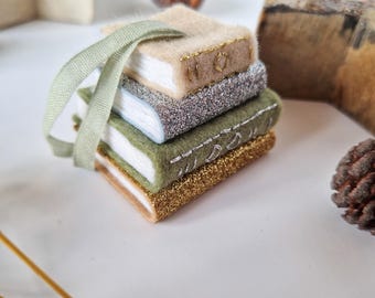 May include: A stack of four miniature book ornaments with white pages. The top book is beige felt with gold stitching, the second is silver glitter, the third is olive green felt with white stitching, and the bottom is gold glitter. A sage green ribbon is attached.