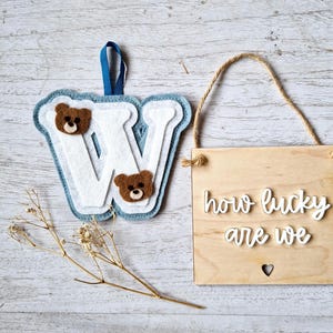 May include: A blue felt letter W with two brown felt bear faces, hanging from a blue ribbon. A wooden sign with the text "how lucky are we" in white lettering is hanging from a twine rope.