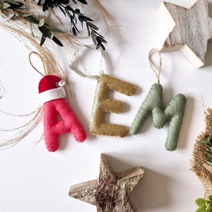Könnte beinhalten: Filzbuchstaben-Ornamente in Rot, Gold und Grün, die das Wort "AEM" buchstabieren. Der rote Buchstabe "A" hat einen weißen Weihnachtsmann-Hut obenauf. Der goldene Buchstabe "E" hat Glitzer. Die Ornamente hängen an einer Schnur und befinden sich auf einem weißen Hintergrund.