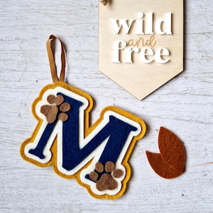 May include: Felt letter M in yellow with a navy blue centre and brown paw prints. The letter is hanging from a brown ribbon. A wooden sign with the text "wild and free" is in the background.