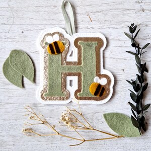 May include: A felt letter H with a gold glitter background and two felt bees with black and yellow stripes. The letter is hanging from a white ribbon.