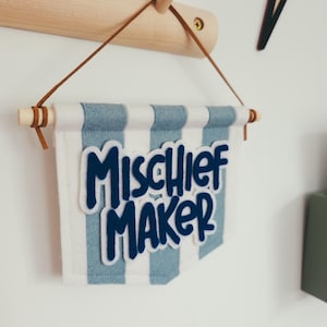 May include: A felt banner with blue and white stripes and the words "Mischief Maker" in dark blue. The banner is suspended by a brown leather cord and wooden dowels. The banner is hanging on a wooden wall hook.