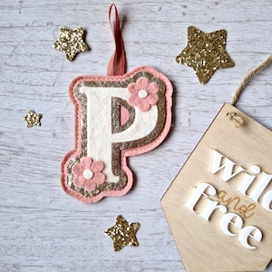 May include: A pink felt letter P with gold glitter accents and a pink felt flower. The letter is hanging from a pink ribbon and is surrounded by gold glitter stars. The text "wild and free" is visible on a wooden sign in the background.