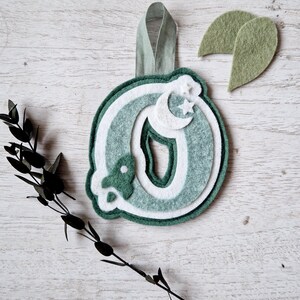 May include: A green felt letter O with a white crescent moon and stars, a green felt leaf, and a green ribbon for hanging.