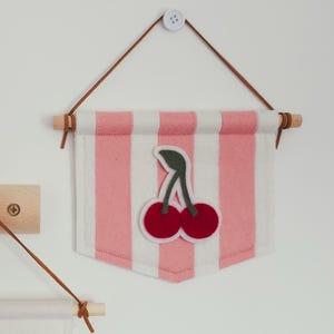 May include: A pink and white striped felt banner with a cherry applique. The banner is suspended by a brown leather cord and wooden dowel. The cherry applique features two red cherries with green stems.