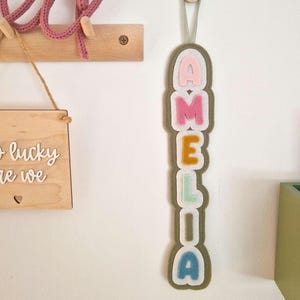 May include: A felt name banner with the name "Amelia" spelled out in colourful letters. Each letter is outlined in white and has a different colour background. The banner is hanging from a white string with a button at the top.