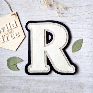 May include: A white felt letter R with a black outline and a beige felt backing. The letter is on a light wood background with green felt leaves.