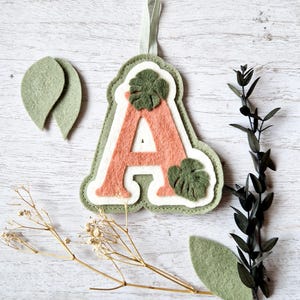 May include: A felt letter A with a pink background and green leaves. The letter is hanging from a white string. There are two green felt leaves on the left side of the image and one on the right side.