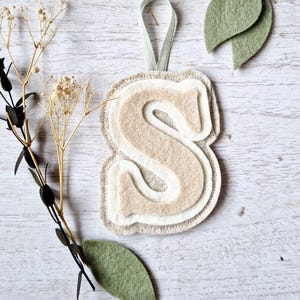 May include: A layered felt letter "S" in beige, cream, and light grey, with a light green ribbon for hanging. Two green felt leaves and a sprig of dried flowers are also present, all on a white wooden surface.