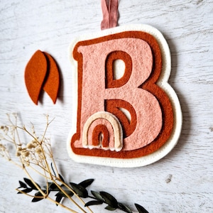 May include: A felt letter B ornament in white, orange, and pink with a small rainbow design. The ornament is hanging from a pink ribbon.