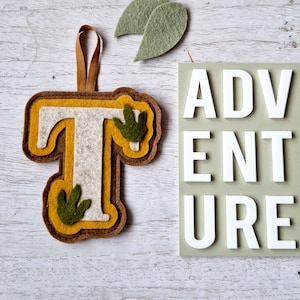 May include: A felt letter T with green leaves, in a brown, yellow, and white colour scheme. The letter is hanging from a brown ribbon. The word "ADVENTURE" is written in white letters on a green background.