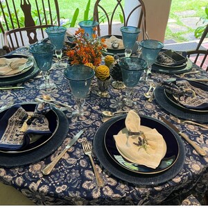Peut inclure: Une table de salle &agrave; manger joliment dress&eacute;e avec une nappe bleue &agrave; motifs floraux, des verres bleus, des assiettes bleu marine et des couverts. Des serviettes pli&eacute;es et une pi&egrave;ce ma&icirc;tresse florale ajoutent une touche de couleur. Chaises en bois.