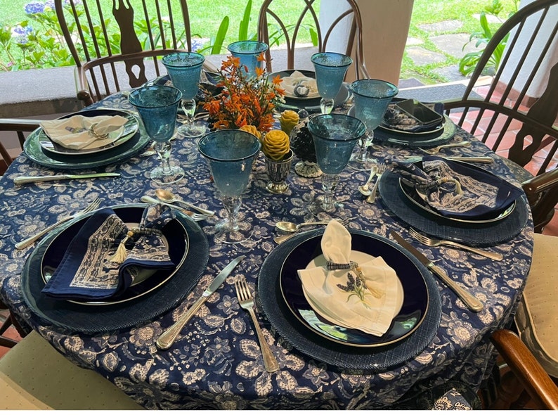 Peut inclure: Une table de salle &agrave; manger joliment dress&eacute;e avec une nappe bleue &agrave; motifs floraux, des verres bleus, des assiettes bleu marine et des couverts. Des serviettes pli&eacute;es et une pi&egrave;ce ma&icirc;tresse florale ajoutent une touche de couleur. Chaises en bois.