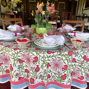 Puede incluir: Un mantel redondo blanco con un patr&oacute;n floral con flores rosas y verdes y frutos de granada rojos. El mantel tiene un borde azul y rosa. Una mesa puesta con vasos rosas y transparentes, servilletas blancas y manteles individuales de mimbre est&aacute; sobre el mantel.