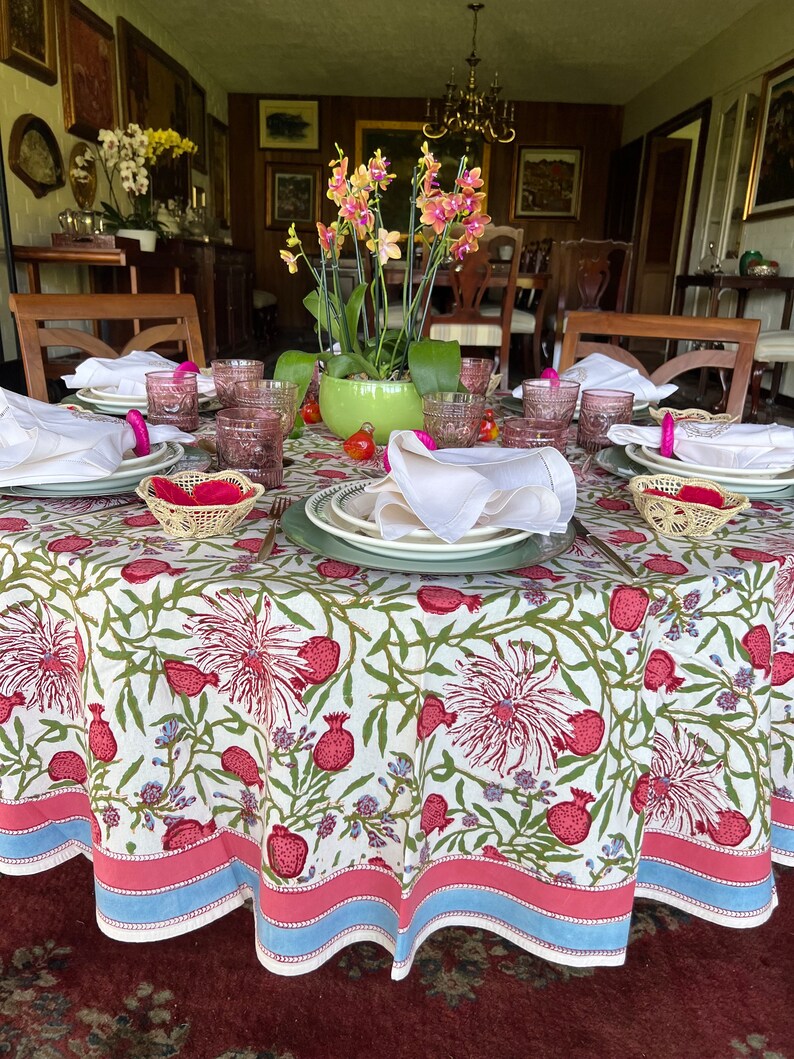 Puede incluir: Un mantel redondo blanco con un patr&oacute;n floral con flores rosas y verdes y frutos de granada rojos. El mantel tiene un borde azul y rosa. Una mesa puesta con vasos rosas y transparentes, servilletas blancas y manteles individuales de mimbre est&aacute; sobre el mantel.